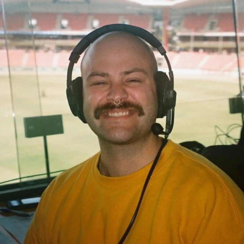 Karl Svikis grinning and wearing a radio headset while presenting at AFTRS Show Radio from a broadcasting booth above the stadium
